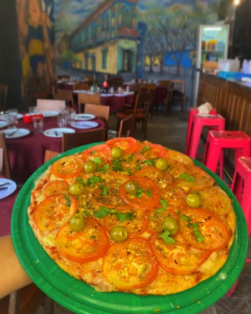 Pizza de tomate y aceitunas frente al salón, con un mural del barrio de La Boca en la pared y banquetas rosas en la barra