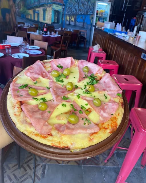 Pizza de muzzarella con jamón y aceitunas en una bandeja, con el salón de Don Sergio detrás